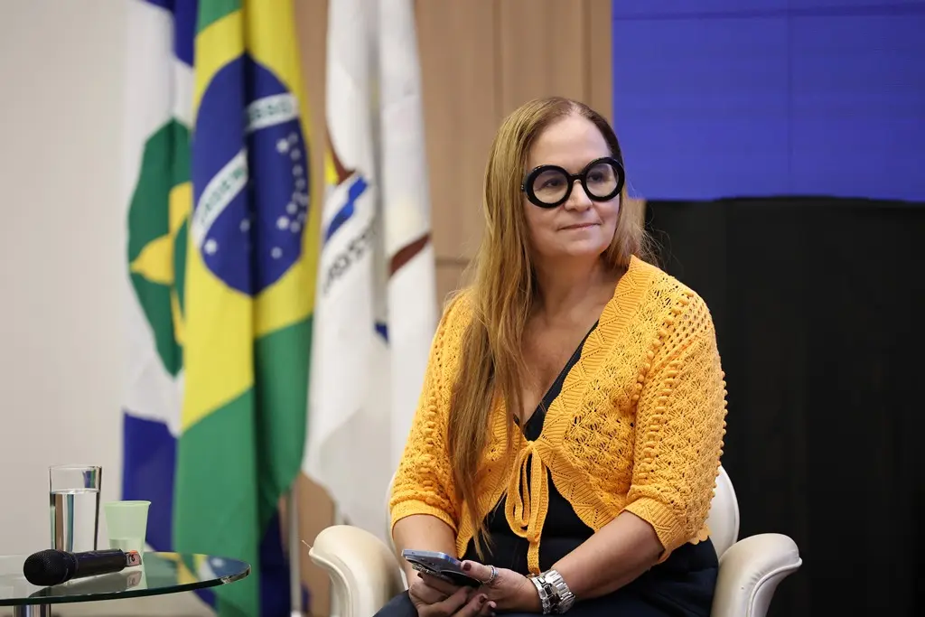 Foto horizontal que mostra a diretora de Fiscalização da ANS, Eliane Aparecida de Castro Medeiros, sentada em uma poltrona. Ela é uma mulher branca, loira, usando vestido preto e boleto amarelo de crochê e óculos de grau de armação preta.
