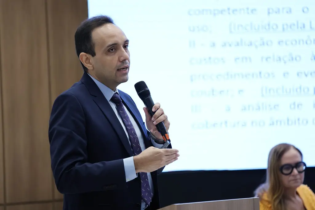 Foto horizontal que mostra o juiz Gerardo Humberto da cintura pra cima, falando ao microfone durante palestra. Ele é um homem pardo, alto, magro, de olhos e cabelos castanhos, usando camisa azul clara, gravata e terno azul marinho.