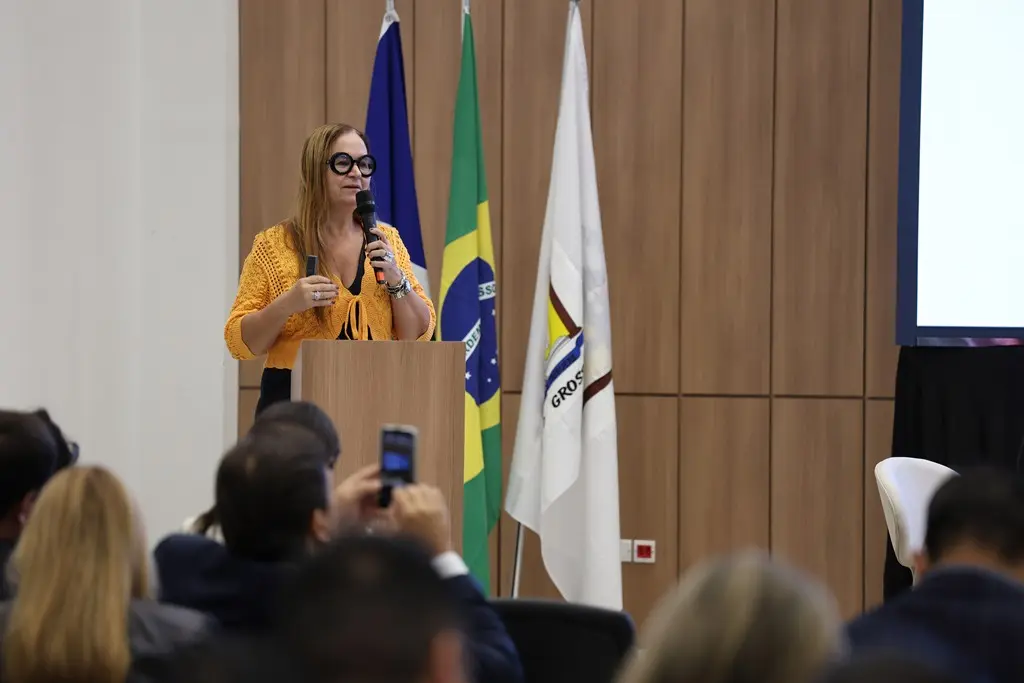 Foto horizontal que mostra a diretora de Fiscalização da ANS, Eliane Aparecida de Castro Medeiros, falando ao microfone em um púlpito. Ela é uma mulher branca, loira, usando vestido preto e boleto amarelo de crochê e óculos de grau de armação preta.