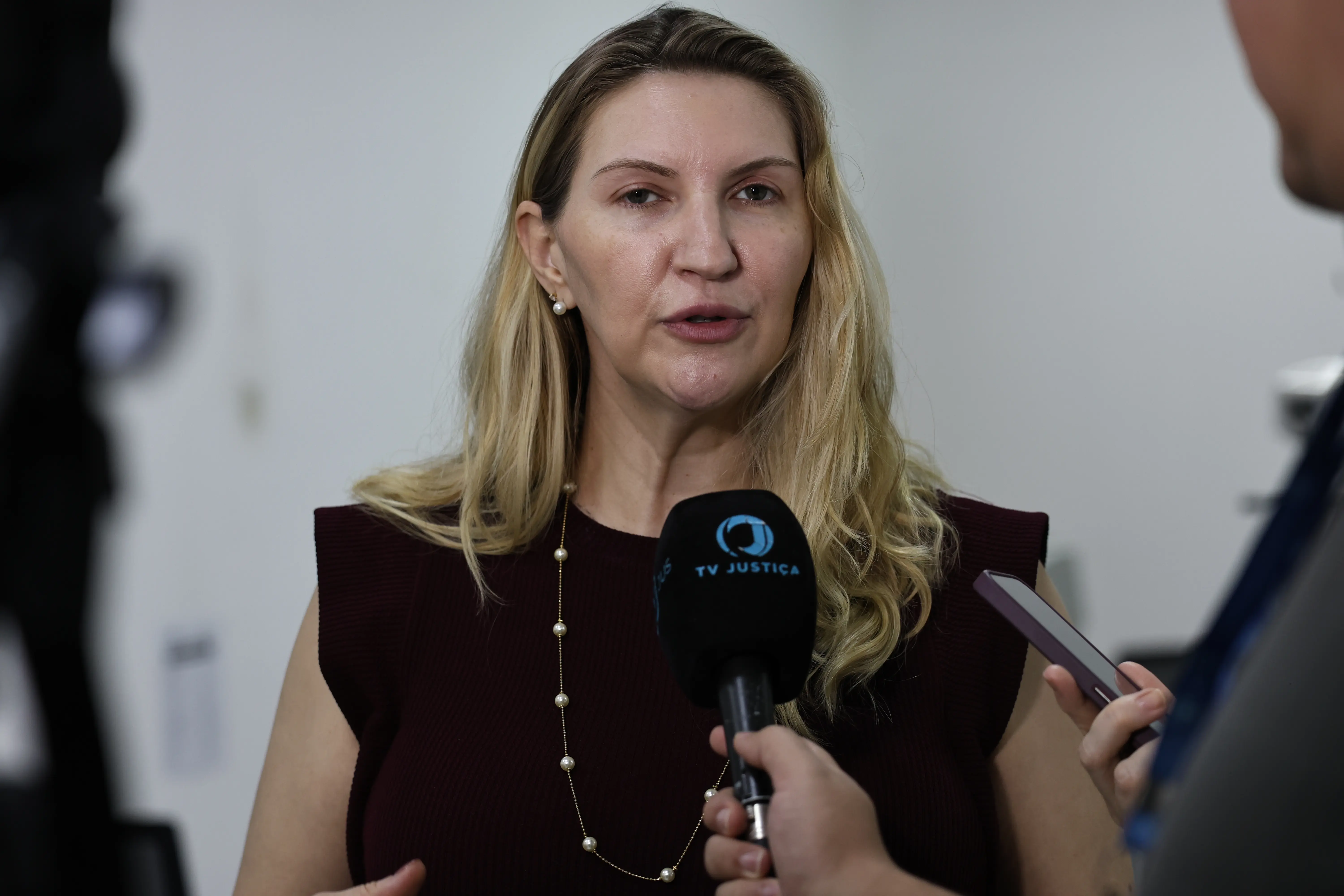 Mulher loira de blusa escura e colar de pérolas fala ao microfone da 