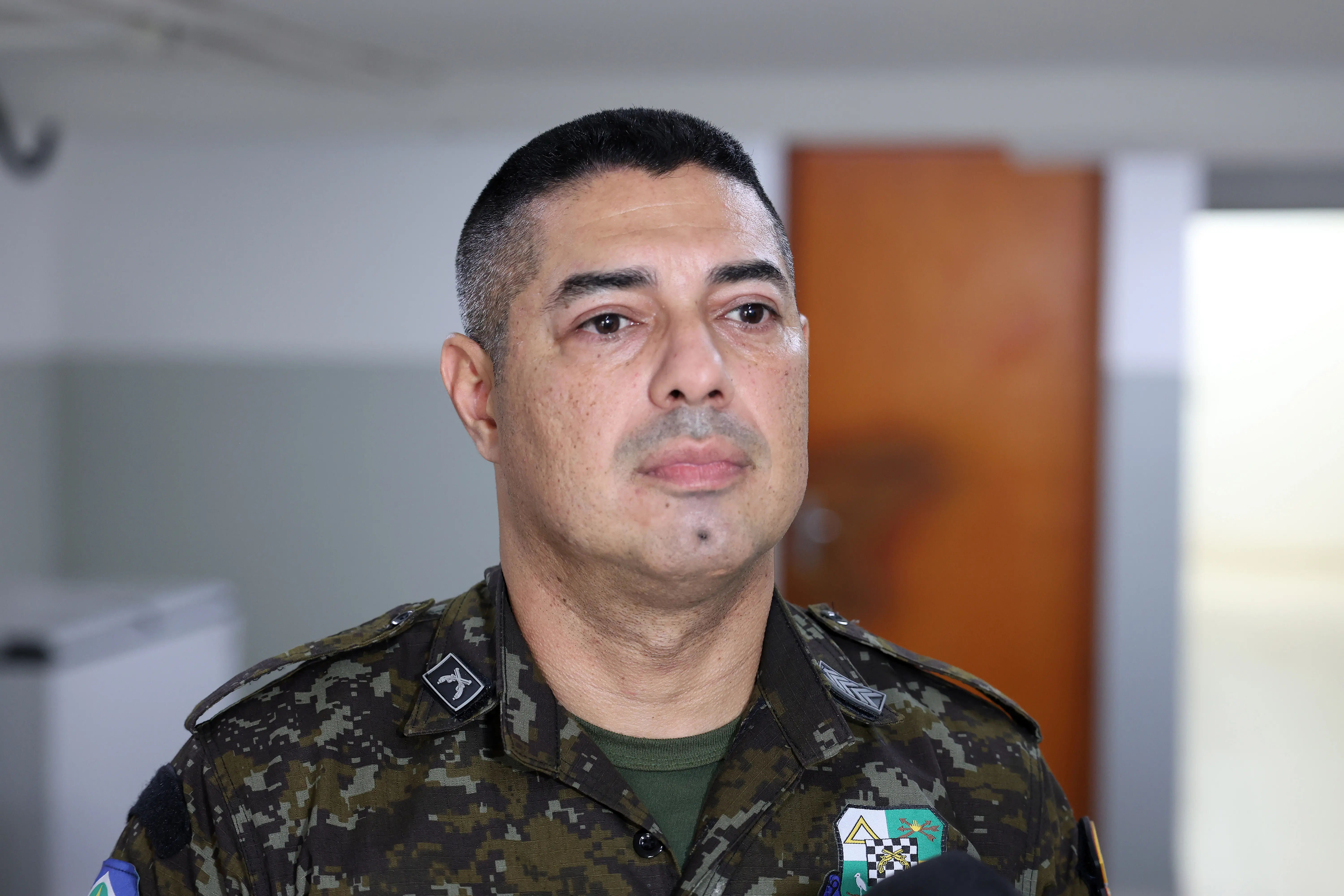 O 1º sargento da Polícia Militar Ambiental e integrante do Juvam, Marcello Amui, aparece em primeiro plano durante entrevista. Ele veste farda camuflada e está em ambiente interno.