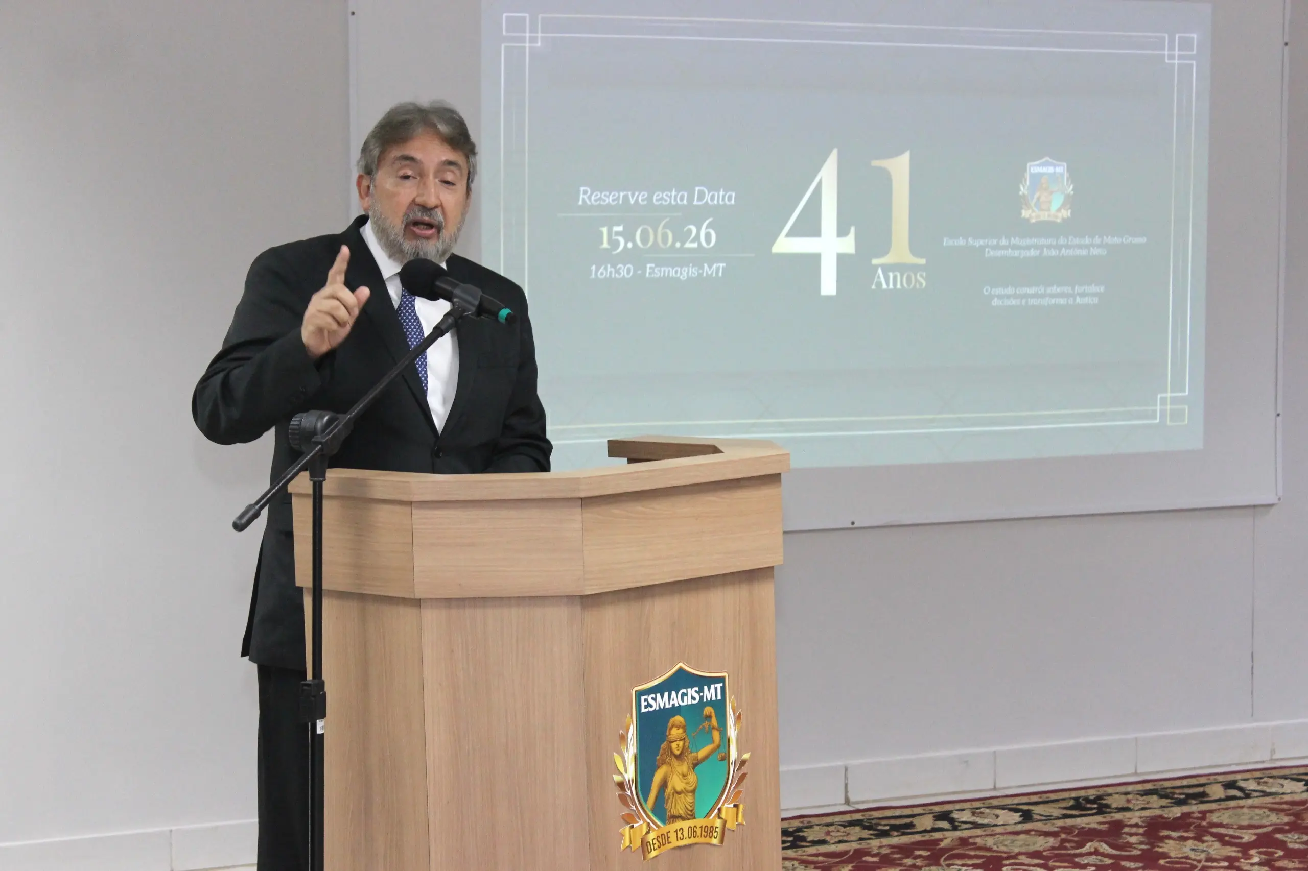 Homem de terno escuro, barba grisalha e gravata azul discursa em um púlpito de madeira clara com o brasão da ESMAGIS-MT fixado na frente. Ele está posicionado à esquerda, com o dedo indicador direito levantado em um gesto enfático diante do microfone. 