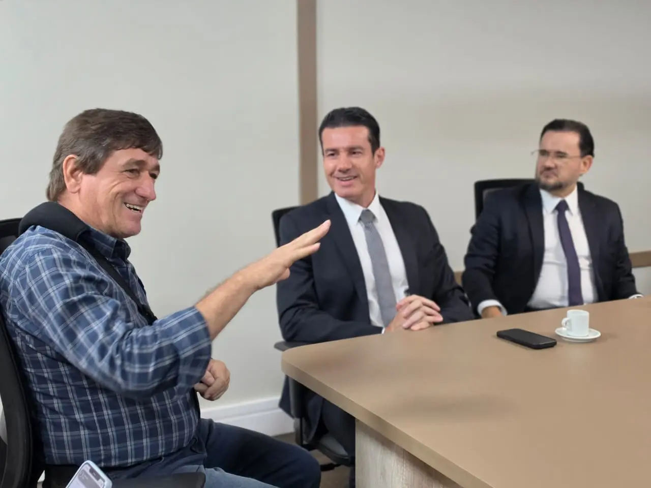 Foto horizontal que mostra o prefeito de Tangará da Serra, o desembargador Rodrigo Curvo e o juiz Bruno D'Oliveira sentados em volta de uma mesa de reunião. O prefeito está na cabeceira sorrindo e gesticulando.
